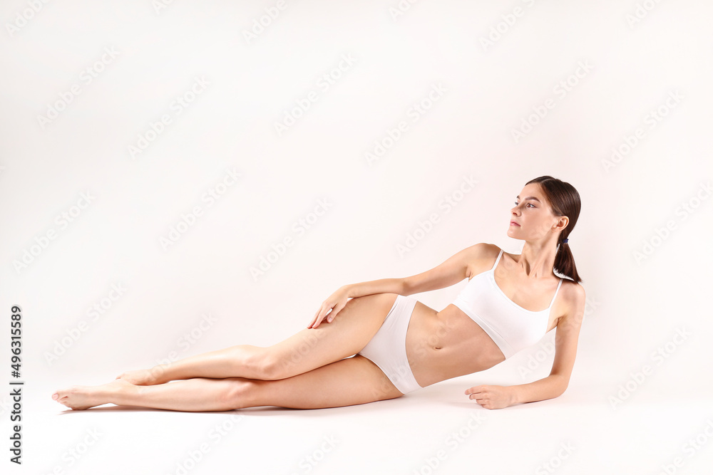 Studio shot of a fit brunette woman with her hair tied in ponytail, wearing seamless underwear, lying on isolated white background. Slim female with flat belly in white lingerie. Copy space for text.