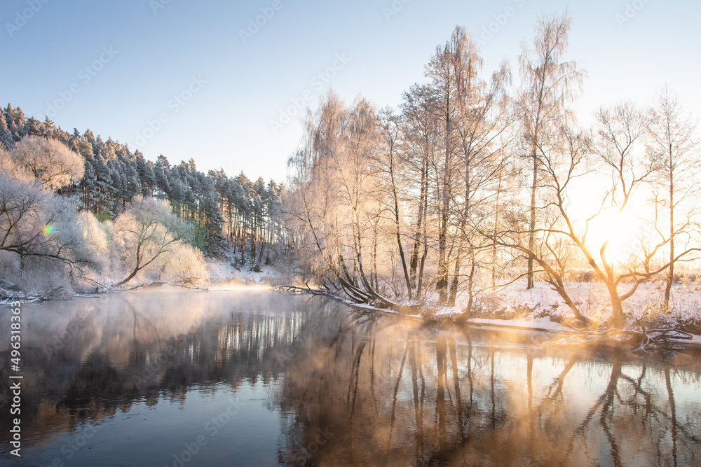 Beautiful morning in winter in the wood. The river without ice in the first month of winter. Bright rays break through small mist and branches of trees.