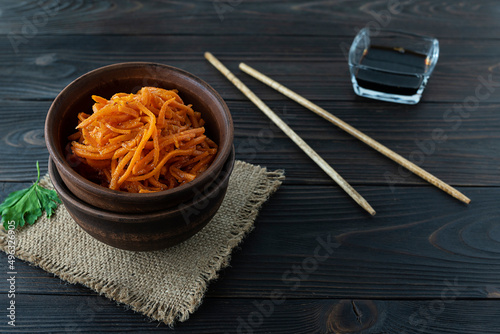 Korean carrot salad in a clay salad bowl on a canvas napkin. Wooden chopsticks on the table. Soy sauce in a glass vase in the background. Dark wooden background