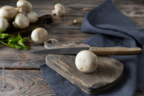 Raw champignon on a brown wooden plank. Knife on the table. Mushrooms and parsley in the background. Wooden background