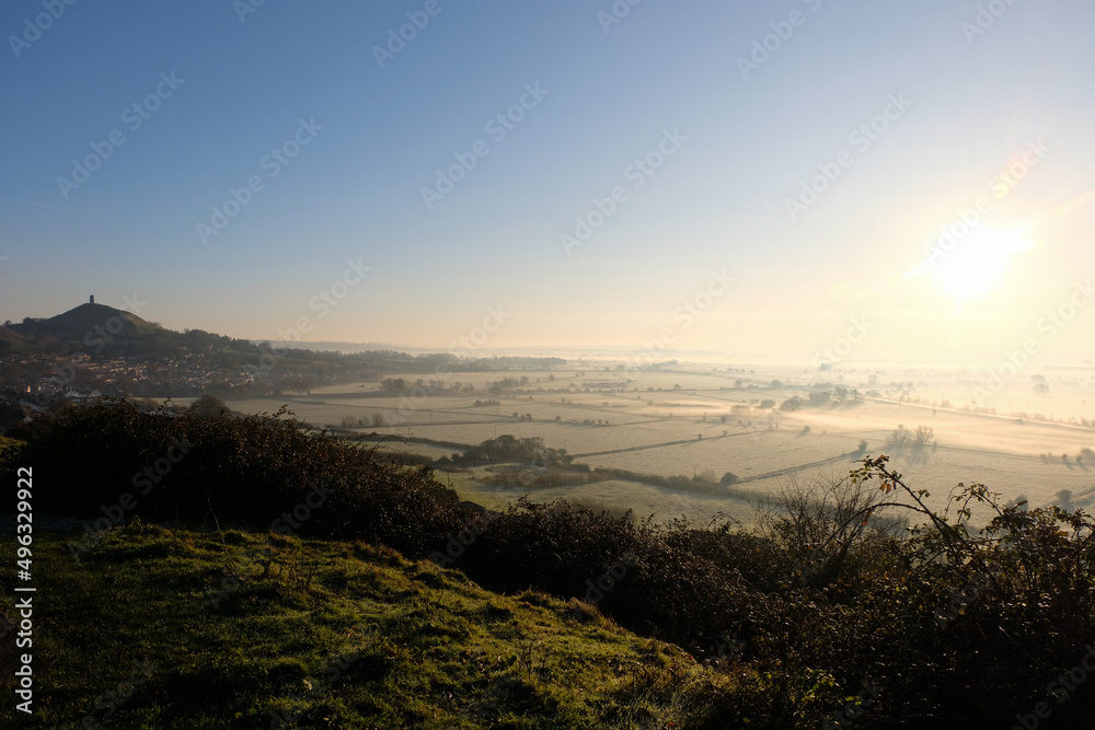 Obraz premium The historic landmark of St Michael's Glastonbury Tor tower overlooking the beautiful rural countryside of Somerset levels with stunning early morning light mist and fog