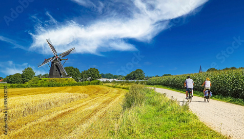 View on cycling path with cyclists couple in rural dutch limburg maas landscape with windmill (Molen de grauwe beer) against blue summer sky - Beesel, Netherlands photo