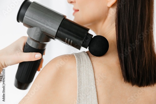 A girl at home massaging with a vibrating massager with a nozzle.  An electric therapeutic pistol massager in her hand massages the muscles of her arm. Sports recovery concept after a workout. 