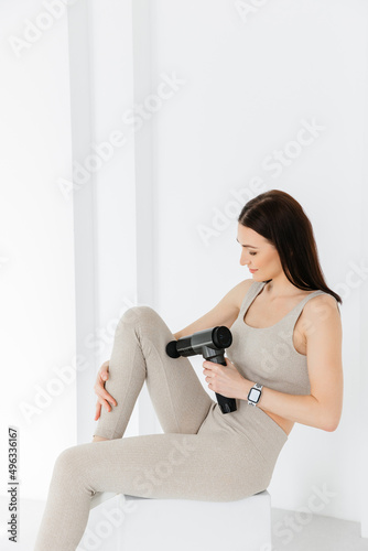 A girl in a tracksuit massaging with a vibrating massager.  An electric therapeutic pistol massager in her hand massages the thigh muscles. Sports recovery concept after a workout. 
