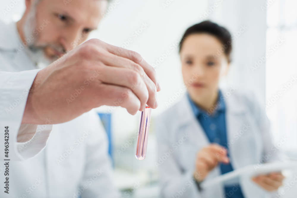 bioengineer holding test tube with liquid while working near blurred colleague. bioengineer holding test tube with liquid while working near blurred colleague.