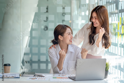 Two young Asian businesswomen successful excited raised hands rejoicing with a laptop computer in a modern office.