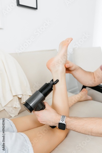 A therapist massages a woman's leg with a massage percussion device in her home. The therapist's hand holds a therapeutic vibrating massager. Physical therapy and muscle recovery and massage