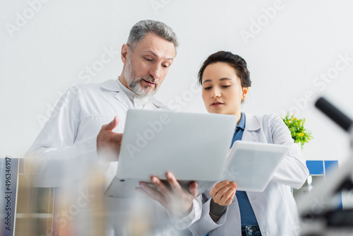 bearded geneticist pointing at laptop near colleague with digital tablet.