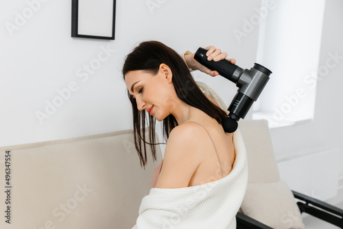 A girl at home holds a therapeutic percussive vibrating massager.  An electric pistol massager in her hand massages her neck muscles. Sports recovery concept after a workout.  photo