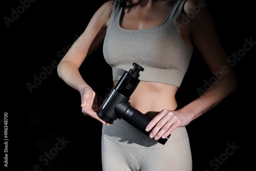 Athletic young woman holding the therapeutic impact massage gun, post-workout recovery. Girl's body in tracksuit against a black background. The nozzles of the vibrating massager. 