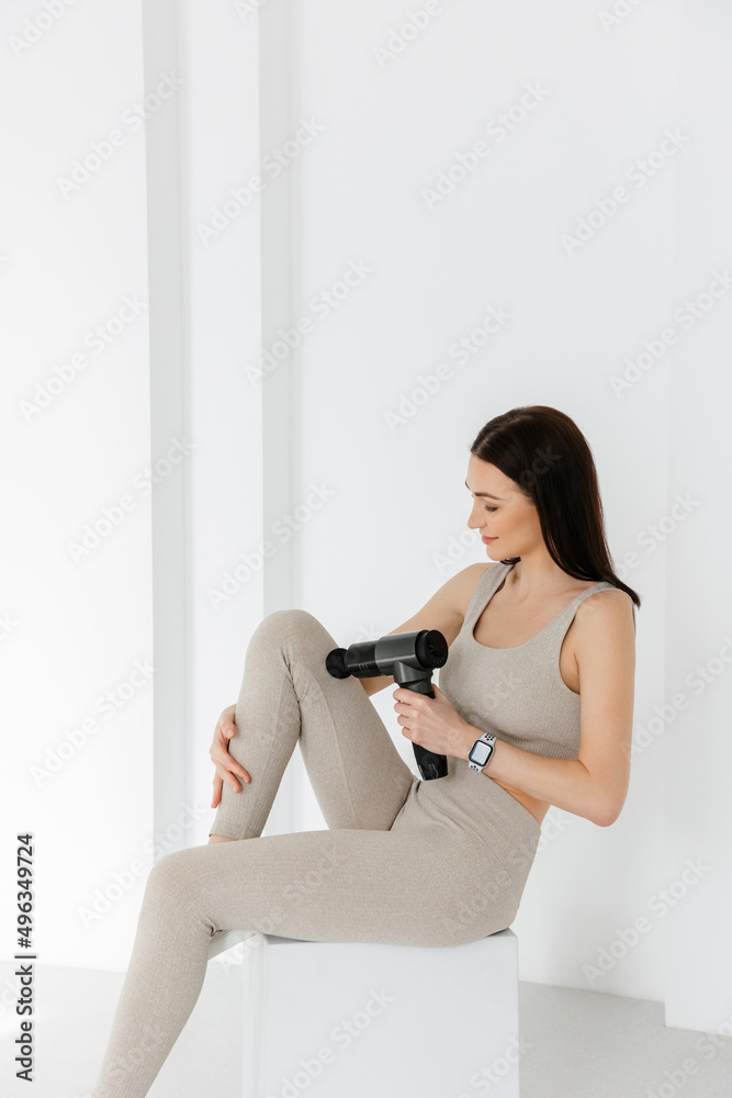 A girl at home on the couch massaging with a vibrating massager. An  electric therapeutic pistol massager in her hand massages her leg muscles.  Sports recovery concept after a workout. Stock Photo