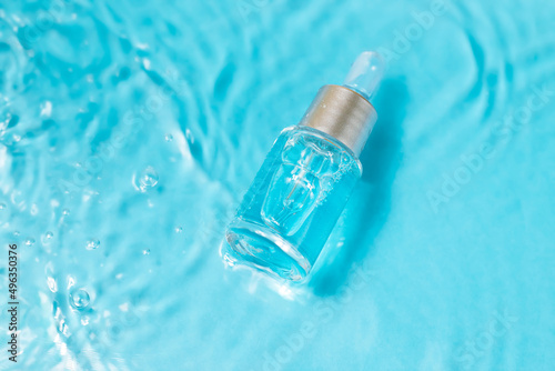 Bottle serum or cosmetic oil in clean transparent water with sunlight on blue background, flat lay, top view, copy space. Cosmetic spa medical skin care concept.