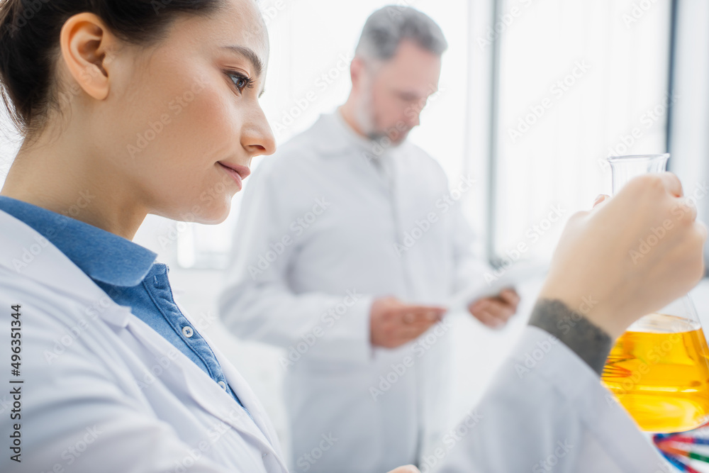 bioengineer holding flask with yellow liquid near colleague on blurred background. bioengineer holding flask with yellow liquid near colleague on blurred background.