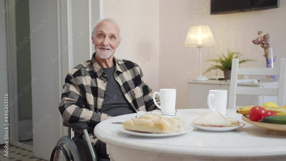 An old man in a wheelchair sits at a dining table and looks at the camera An old man in a wheelchair sits at a dining table and looks at the camera