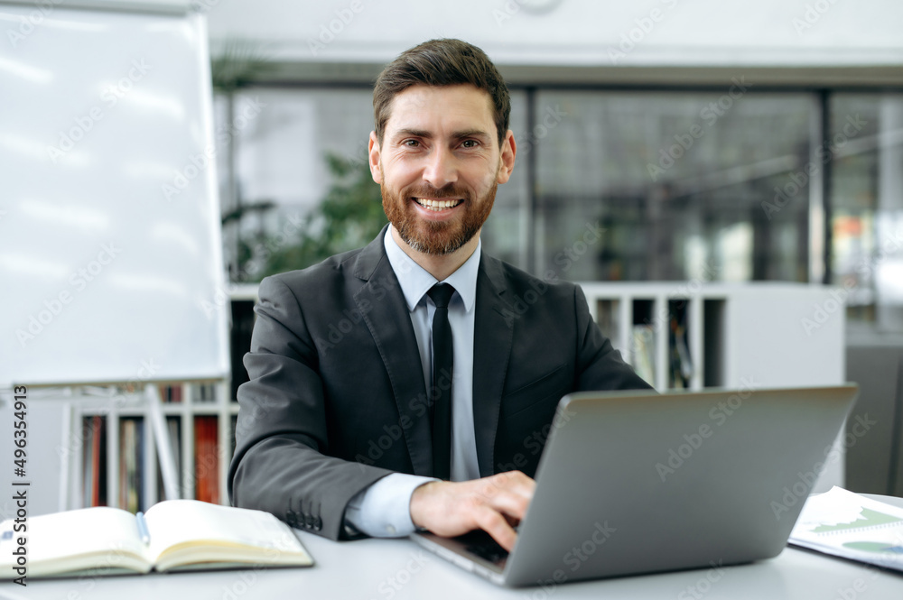 Portrait of a successful attractive Caucasian businessman, tester, IT specialist, sitting at a desktop in front of a laptop, in modern office, in stylish business clothes, looking at camera, smiles