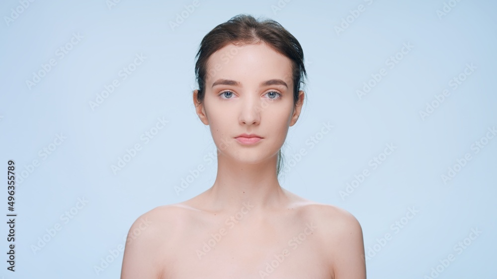 Obraz premium Close-up beauty portrait of young brunette woman standing in front of the camera looks at the camera and smiles against a blue background | Skin care product commercial concept