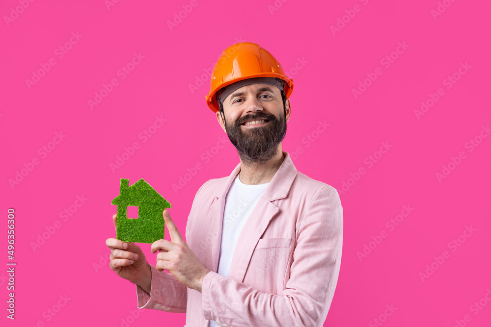 Portrait of young construction engineer wear orange hard hat, in a pink jacket standing on red studio background. A man holds a green eco house. Portrait of young construction engineer wear orange hard hat, in a pink jacket standing on red studio background. A man holds a green eco house.