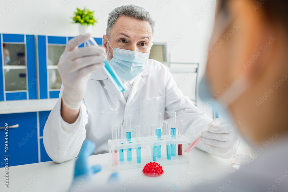 bioengineer in medical mask holding test tube near blurred colleague and coronavirus bacteria model. bioengineer in medical mask holding test tube near blurred colleague and coronavirus bacteria model.