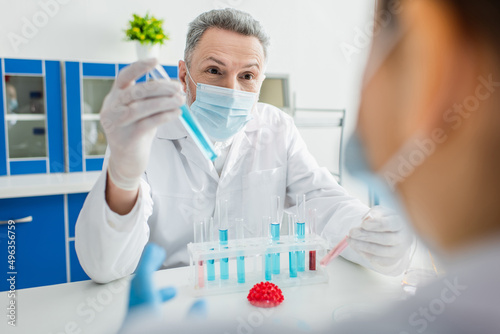 Fototapeta Naklejka Na Ścianę i Meble -  bioengineer in medical mask holding test tube near blurred colleague and coronavirus bacteria model.