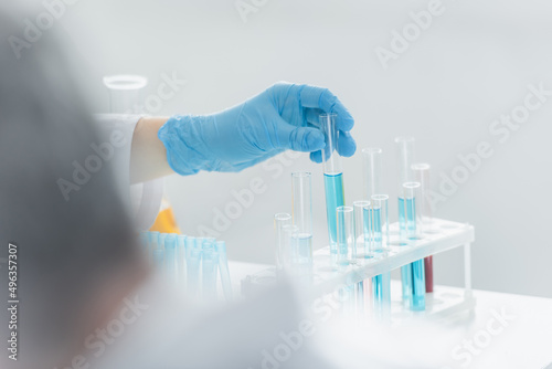 partial view of geneticist holding test tube with blue liquid on blurred foreground.