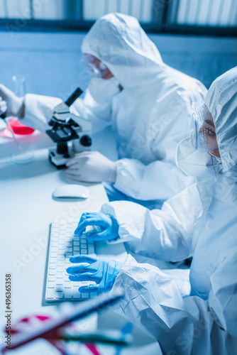woman in hazmat suit typing on computer keyboard near blurred scientist with microscope.