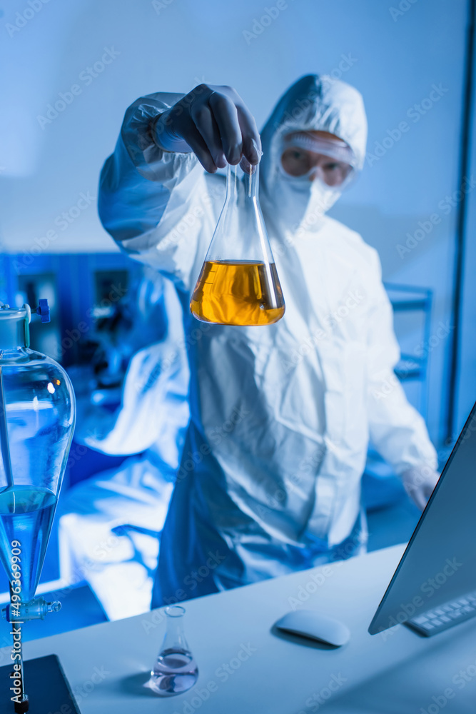 blurred scientist in hazmat suit holding flask with orange liquid in laboratory. blurred scientist in hazmat suit holding flask with orange liquid in laboratory.