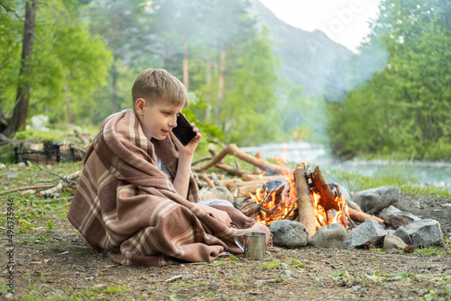happy teenage boy using phone sitting near fire on camping on mountain