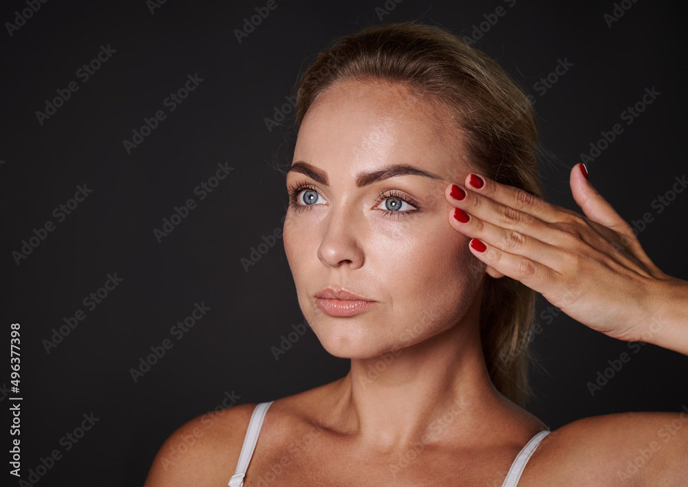 Close-up beauty portrait of a beautiful 30 years old blonde woman with glowing fresh healthy skin holding her hand on her temple, isolated over black background with copy space Close-up beauty portrait of a beautiful 30 years old blonde woman with glowing fresh healthy skin holding her hand on her temple, isolated over black background with copy space