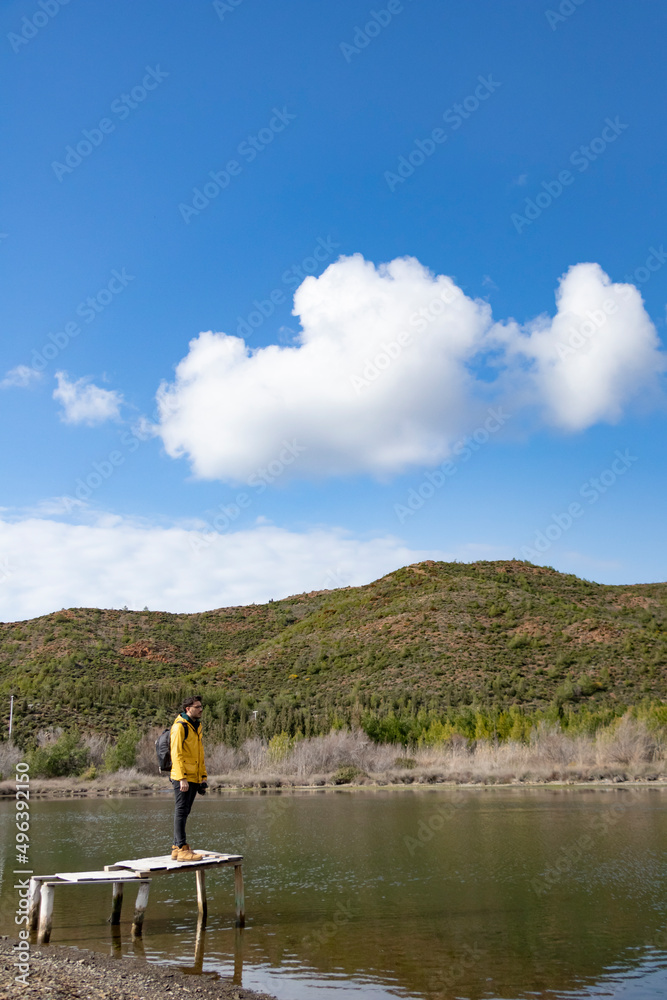 Young, backpacker, traveler boy on old small dock above sea. He is looking beautiful river and mountain view. He has yellow raincoat. Young, backpacker, traveler boy on old small dock above sea. He is looking beautiful river and mountain view. He has yellow raincoat.