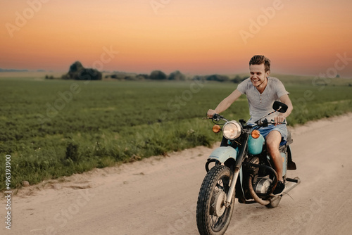 young joyful man rides a retro motorcycle on a country road. The concept of a biker in his youth in the countryside.
