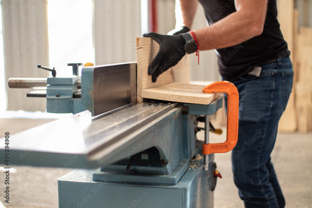 hands of a young carpenter working on an Electric Benchtop Jointer machine cutting a wooden board in a carpentry workshop using safety black gloves hands of a young carpenter working on an Electric Benchtop Jointer machine cutting a wooden board in a carpentry workshop using safety black gloves