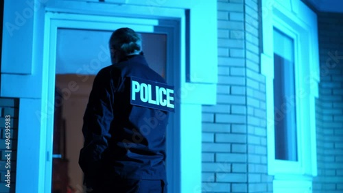 Back view patrolman in uniform standing in front of dark house talking on phone as police lights flashing. Medium shot of Caucasian policeman at crime scene outdoors