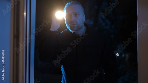 Medium shot of concentrated Caucasian patrolman with flashlight looking inside dark house at night through window. Front view portrait of serious policeman at crime scene outdoors