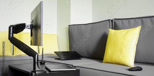 Swivel arm with gas lift holds a modern computer monitor on the table. Next to a laptop, an electronic tablet, a stylus, a computer mouse and a large loft sofa with yellow pillows photo