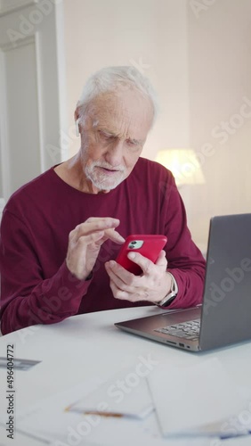 An elderly man sits at a table in the living room and flips through photos on a smartphone