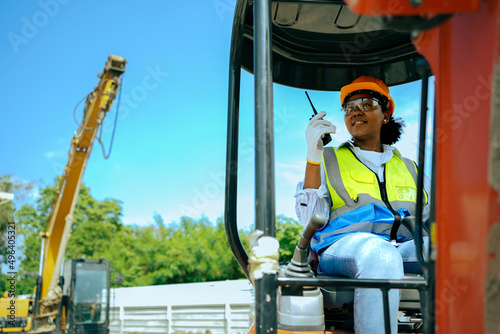 A woman worker driving a backhoe to dig a hole in a construction site holding a walkie talkie.African American female engineer wearing a hard hat and vest.cute female with black skin gender equality.