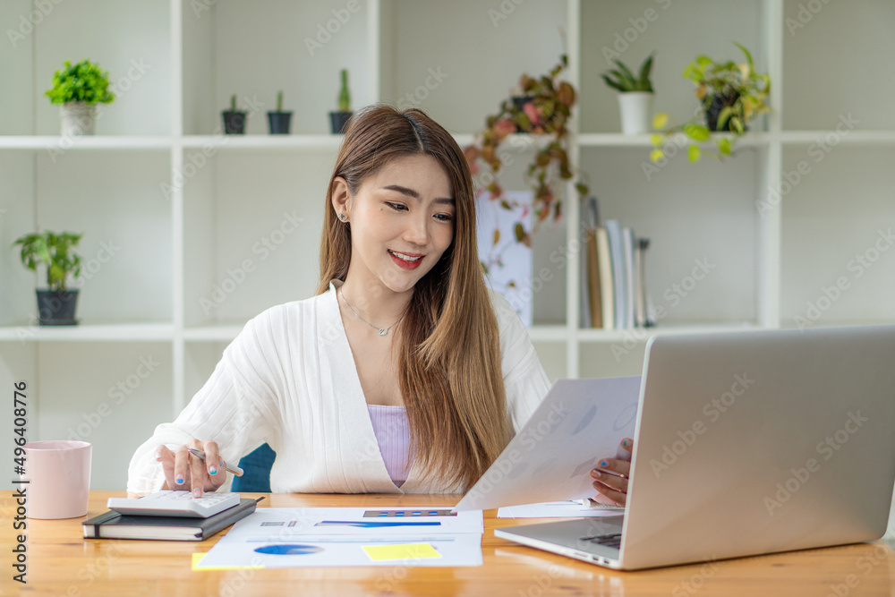 Asian female accountant or banker calculating savings, finance, and economy concepts via laptop. Businesswoman writing charts and graphs that show results in computational analysis.
