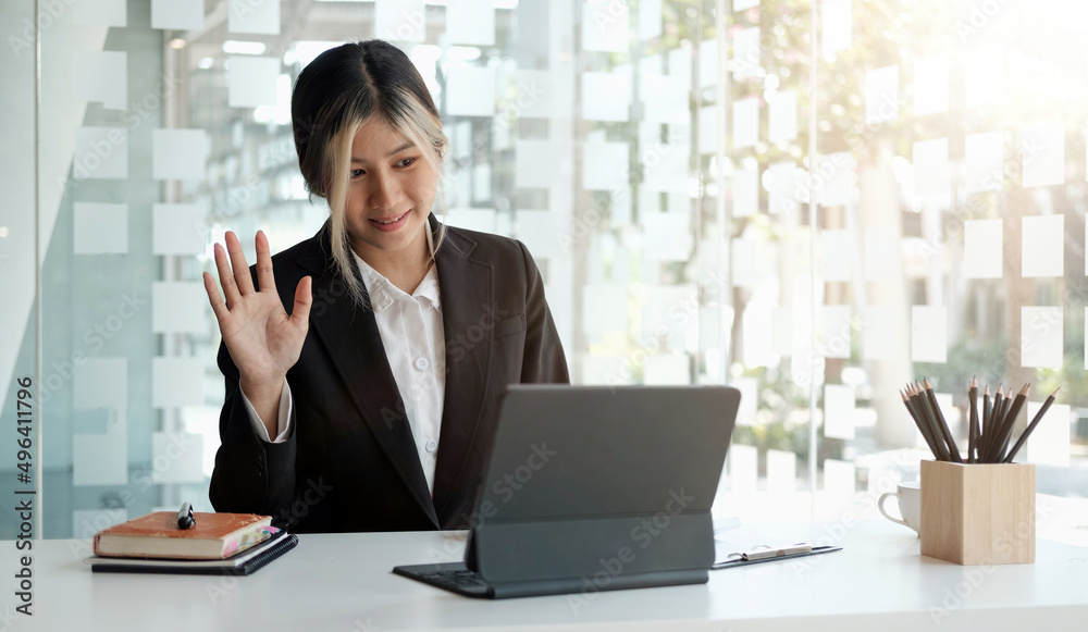 Young asian woman wearing suit smiling and waving hand at laptop, while speaking or chatting on video call in office. Young asian woman wearing suit smiling and waving hand at laptop, while speaking or chatting on video call in office.