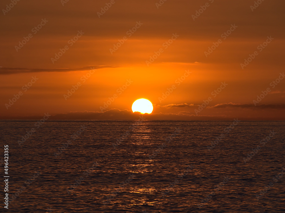 Sunset above the Atlantic Ocean with bright sun disappearing between low clouds on the horizon and orange sky viewed from the coast of Gran Canaria.