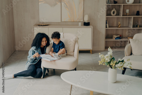 Little mixed race boy reading with his loving mom while enjoying time togerher in living room at home photo