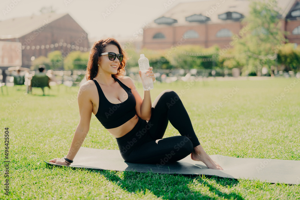 Fototapeta premium Woman has rest after training poses on fitness mat with bare feet drinks water dressed in activewear sunglasses leads healthy lifestyle