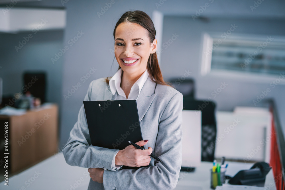 A busy businesswoman standing with clipboard in hands at work and smiling at the camera. A busy businesswoman standing with clipboard in hands at work and smiling at the camera.