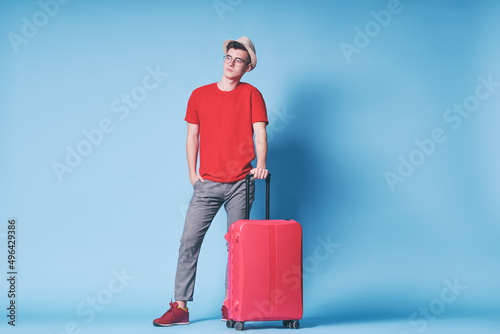 Travel concept. Full length colorful studio portrait of young man in hat with valise on blue background.