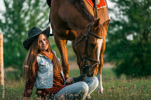 A cowgirl sits on the ground near her horse in a field in summer.