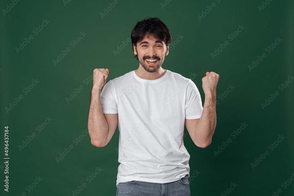 On a dark green background, a young man in a white T-shirt rejoices with a victorious expression