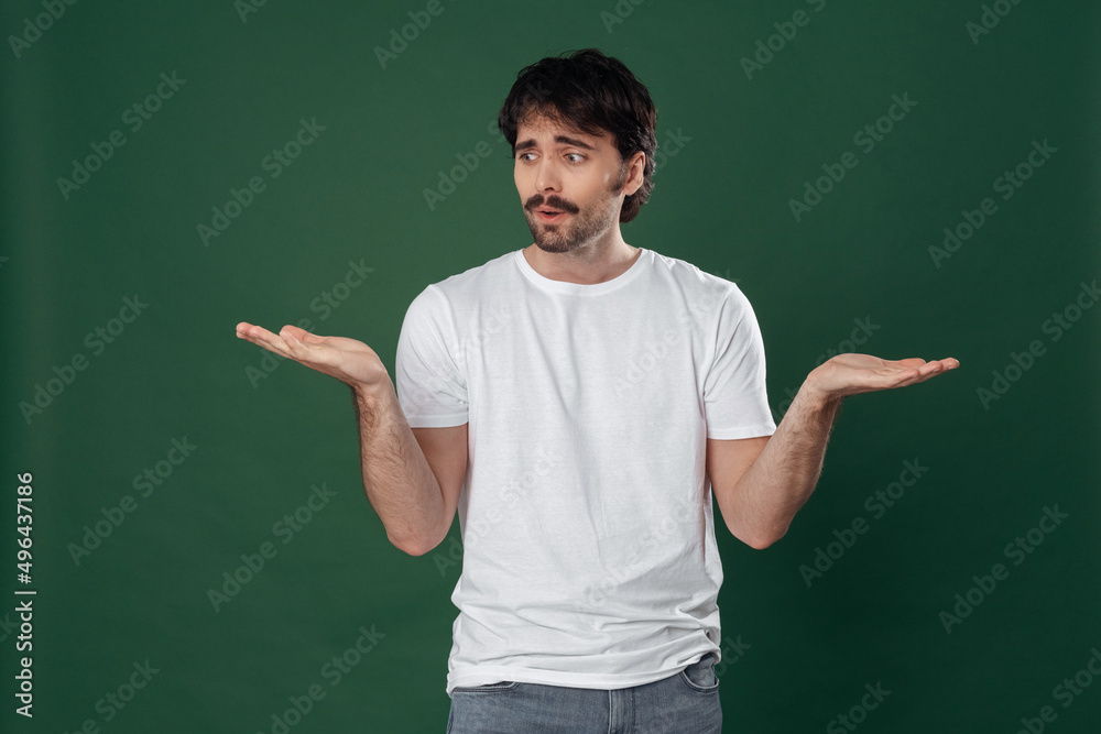 On a dark green background, a young man in a white T-shirt spread his arms to the sides and looks at his left hand On a dark green background, a young man in a white T-shirt spread his arms to the sides and looks at his left hand