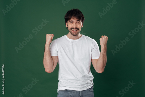 On a dark green background, a young man in a white T-shirt rejoices with a victorious expression © Оlgart