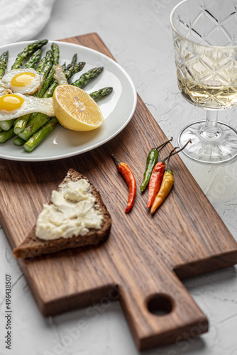 On a wooden board is a plate with scrambled eggs, asparagus and olives. Nearby lies a toast with soft cheese