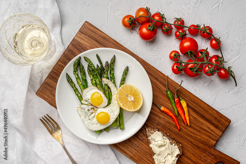 On a gray background  a white plate with scrambled eggs and asparagus stands on a wooden board. View from above. Nearby is a glass of white wine  fresh tomatoes  hot peppers and toast with homemade ch