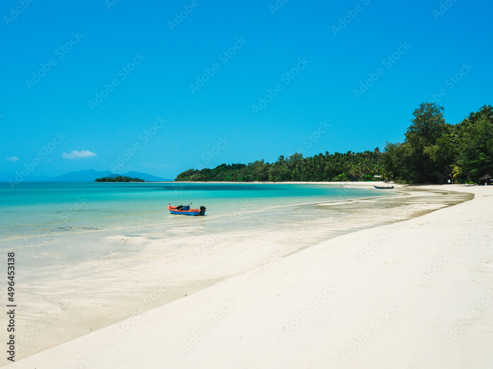 Scenic view of Koh Mak Island peaceful white sand beach with crystal clear turquoise water and local fishing boat against clear blue sky. Trat, Thailand.